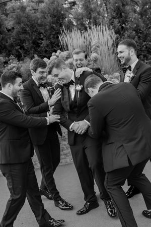 Groomsmen in black suits playfully huddle together at an outdoor wedding celebration.