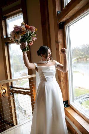 Bride in off-shoulder white gown celebrating with colorful bouquet raised by large windows overlooking water.
