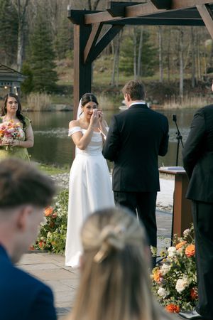 Emotional bride wiping tears during outdoor wedding ceremony by a lake, with floral arrangements and a wooden arbor.