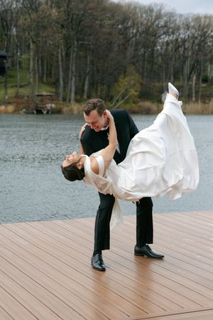 Groom dipping bride in white wedding dress on wooden dock by lake, sharing romantic dance outdoors.