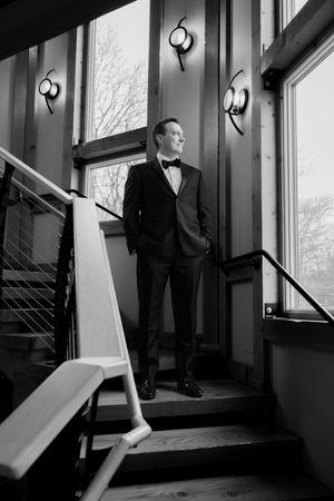 Black and white photo of a groom in a tuxedo standing on stairs by large windows, looking thoughtfully to the side.