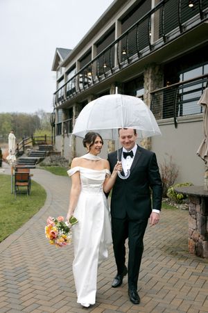 Bride and groom walk under clear umbrella on rainy wedding day outside elegant venue, bride holding colorful bouquet.