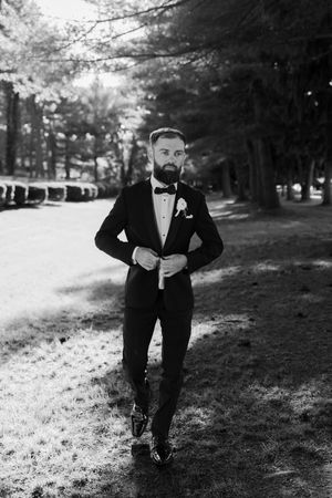 A bearded groom in a black tuxedo and bow tie adjusts his jacket while standing in a tree-lined path.