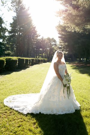 Bride in white lace ballgown with veil holds bouquet on sunlit green lawn surrounded by tall trees.