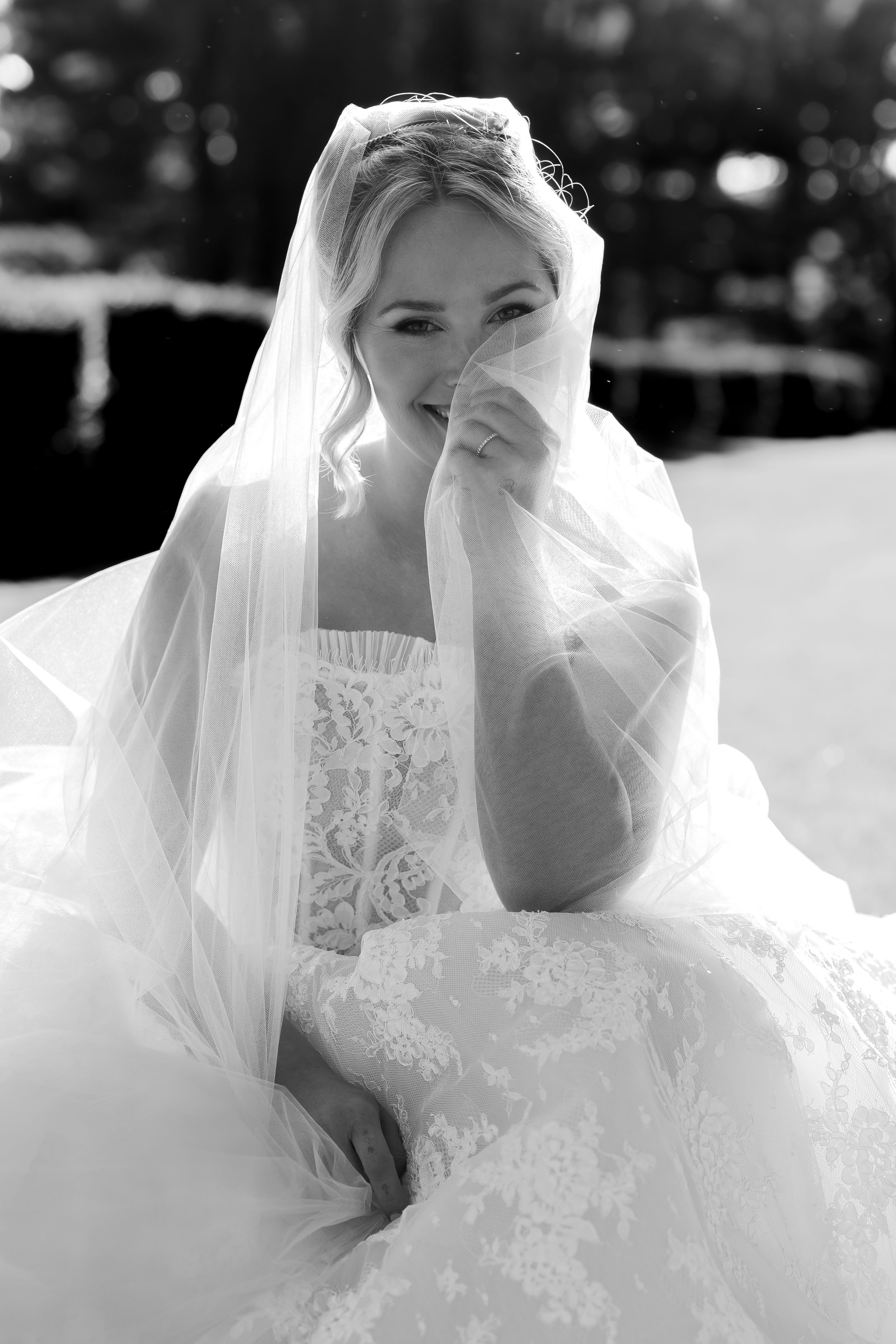 Black and white photo of a bride in lace gown holding her veil near her face while seated outdoors.