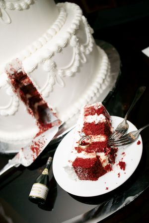 Red velvet wedding cake slice on white plate beside champagne bottle, with two-tier white frosted cake in background.