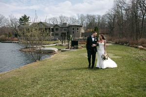 A bride and groom walk hand in hand on a lush green lawn beside a serene lake with an elegant stone mansion backdrop.