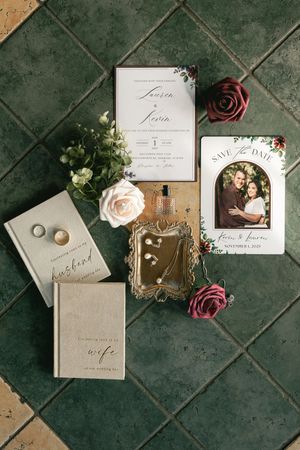 Wedding stationery flat lay with rings, roses, and calligraphy invitations on green tile background.