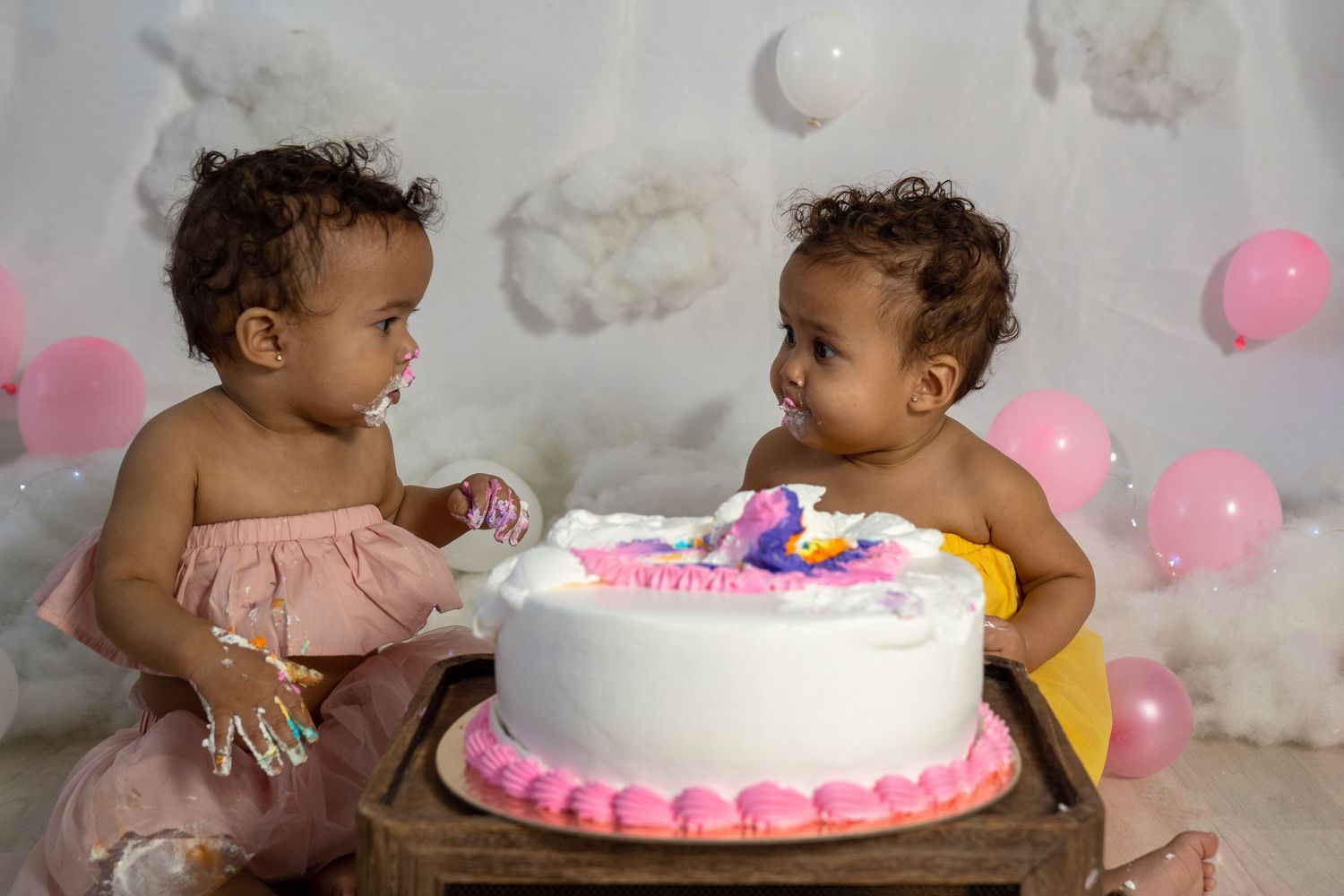 Two toddlers share a pink birthday cake decorated with cloud and moon backdrop.