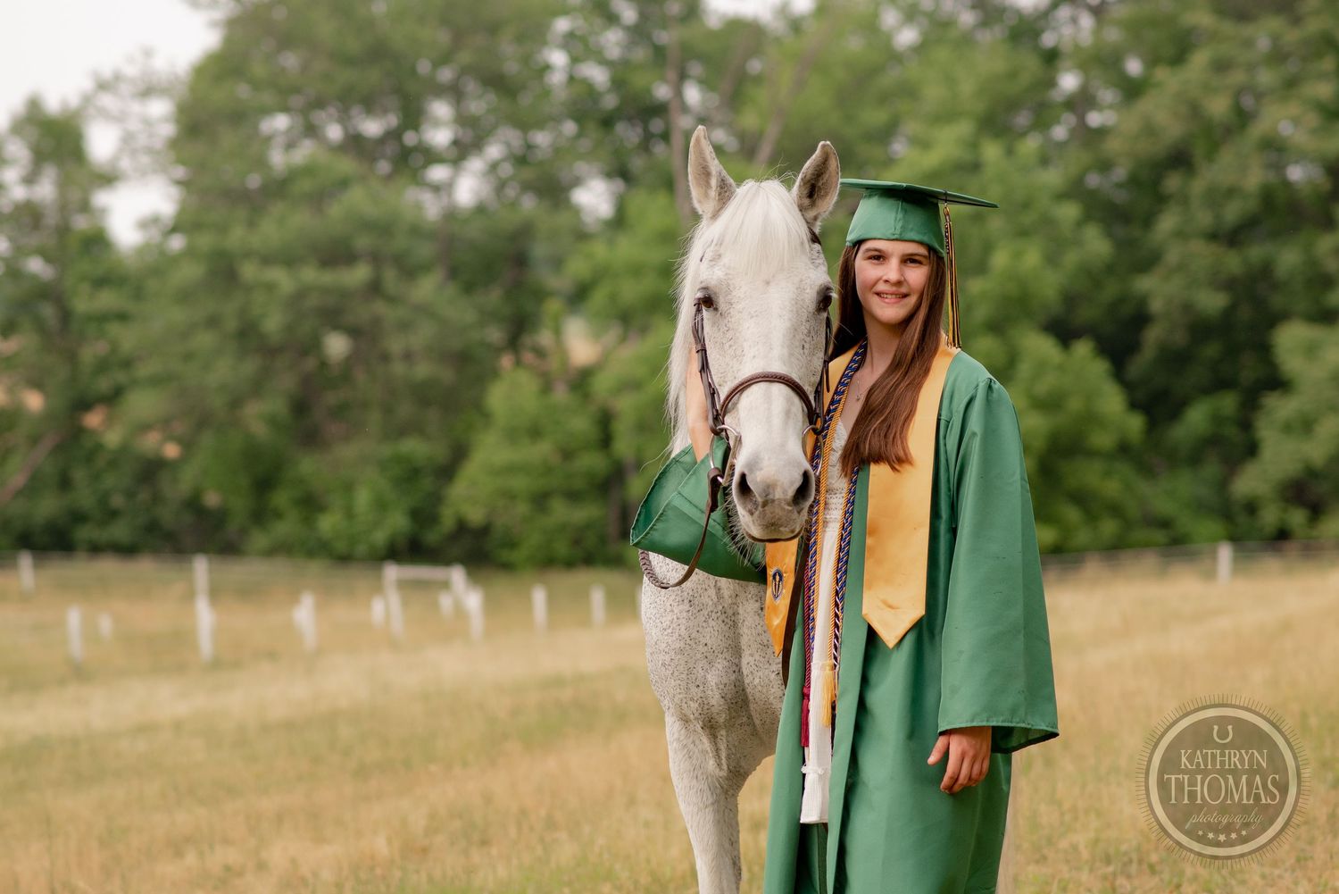 Graduate in green commencement attire stands with friendly white horse in summer field.