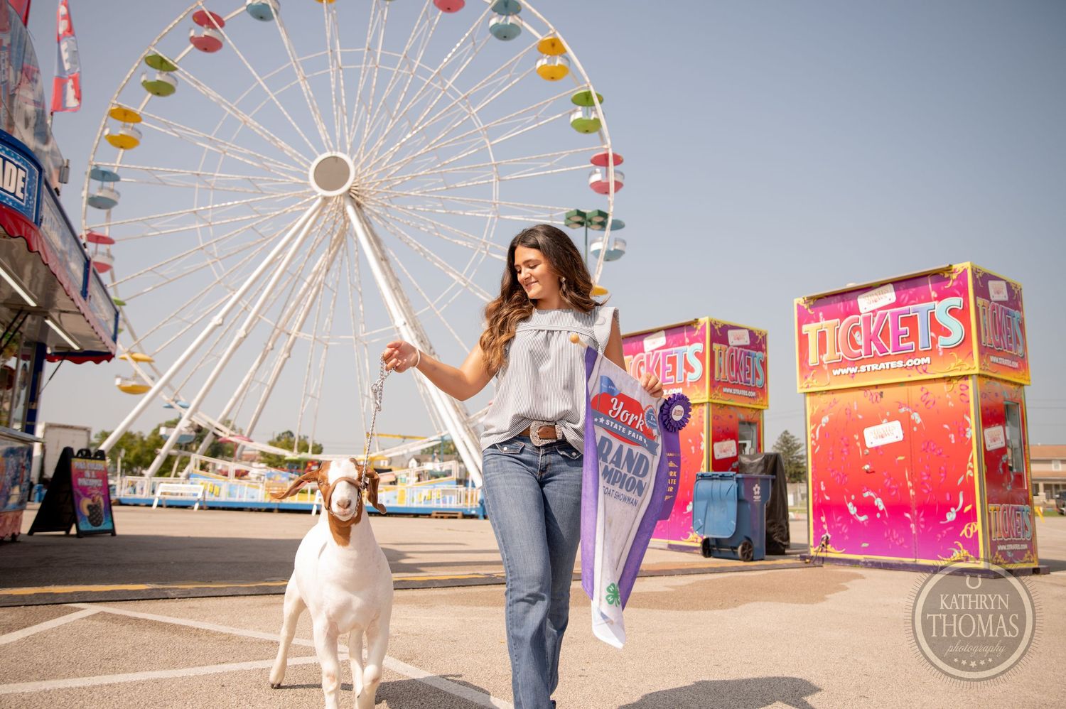 A white dog and companion walking past a vibrant ferris wheel at a carnival.