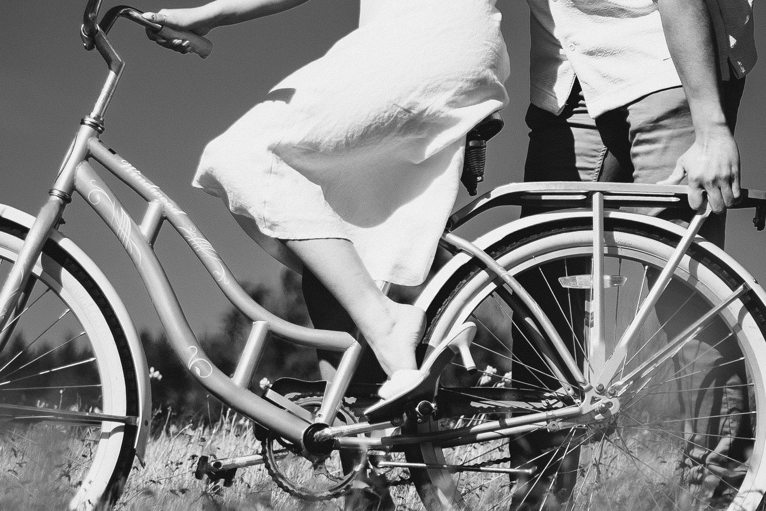 Natalie in heels and a dress sitting on a bike with her fiance holding it steady, in black and white.
