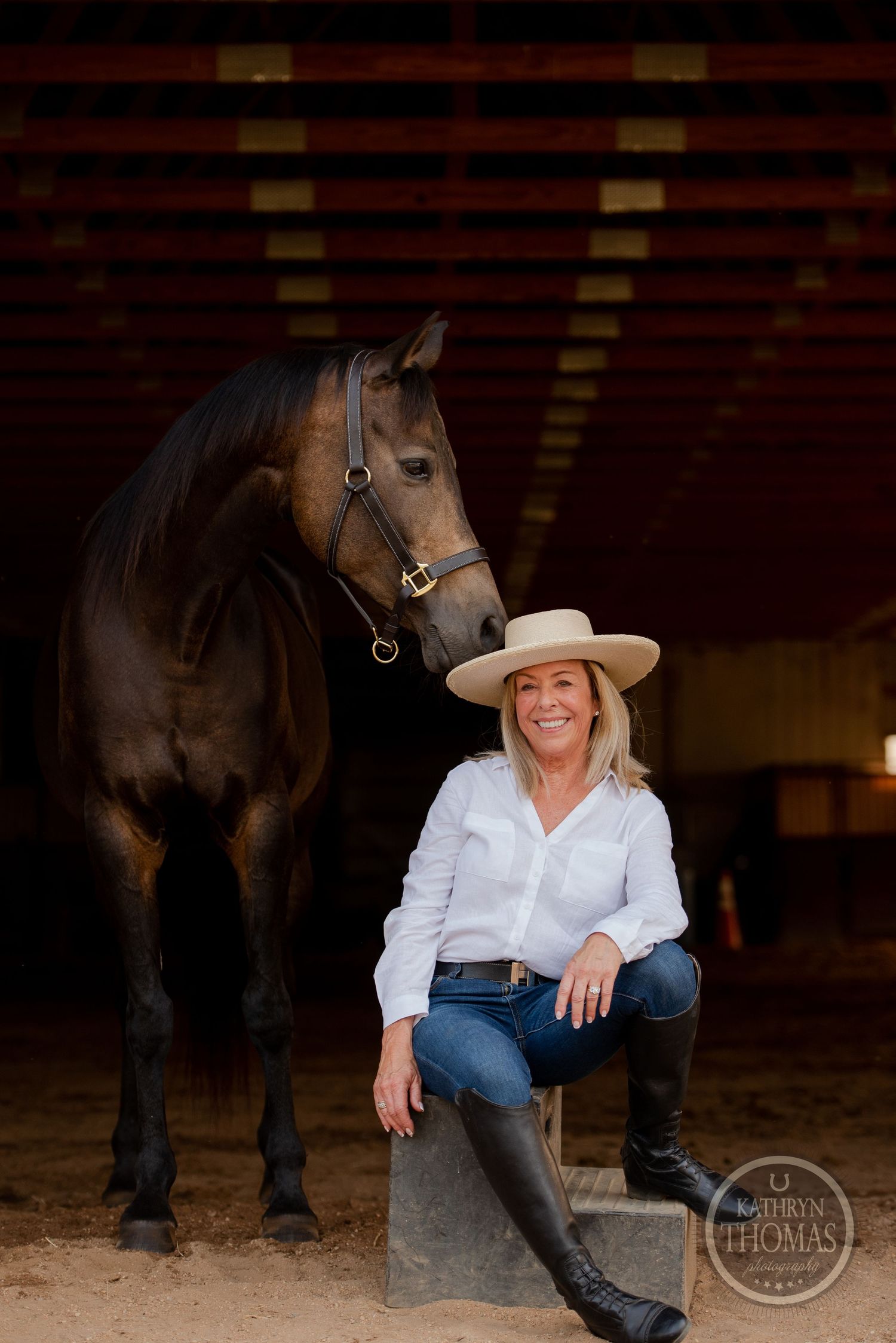 Rider in western wear sits on block beside tall dark horse in indoor arena setting.