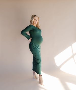 Stylish maternity photo in a long green dress with dramatic window lighting.
