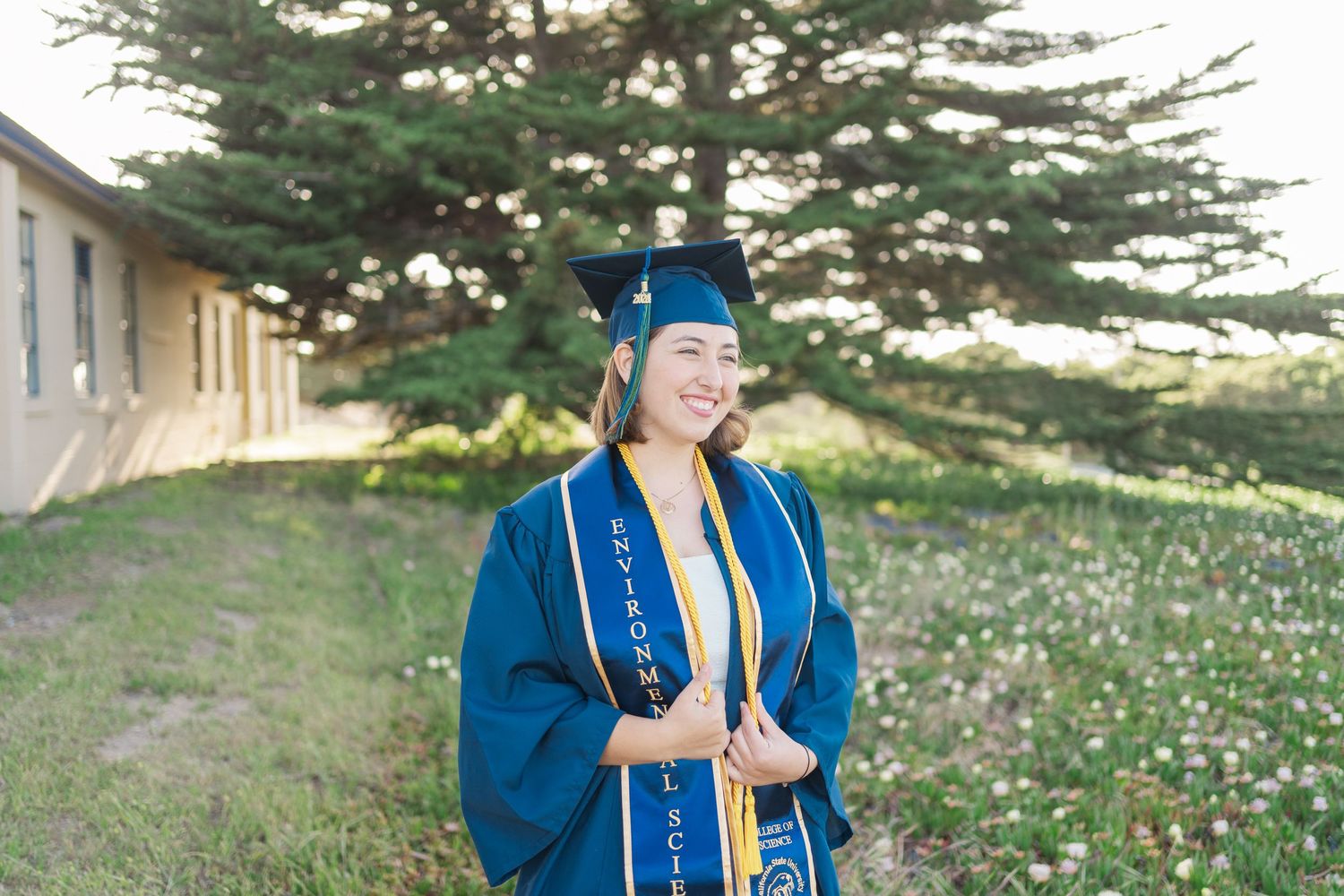 Graduate in blue cap and gown stands smiling outdoors on grassy hillside with evergreen tree in background.