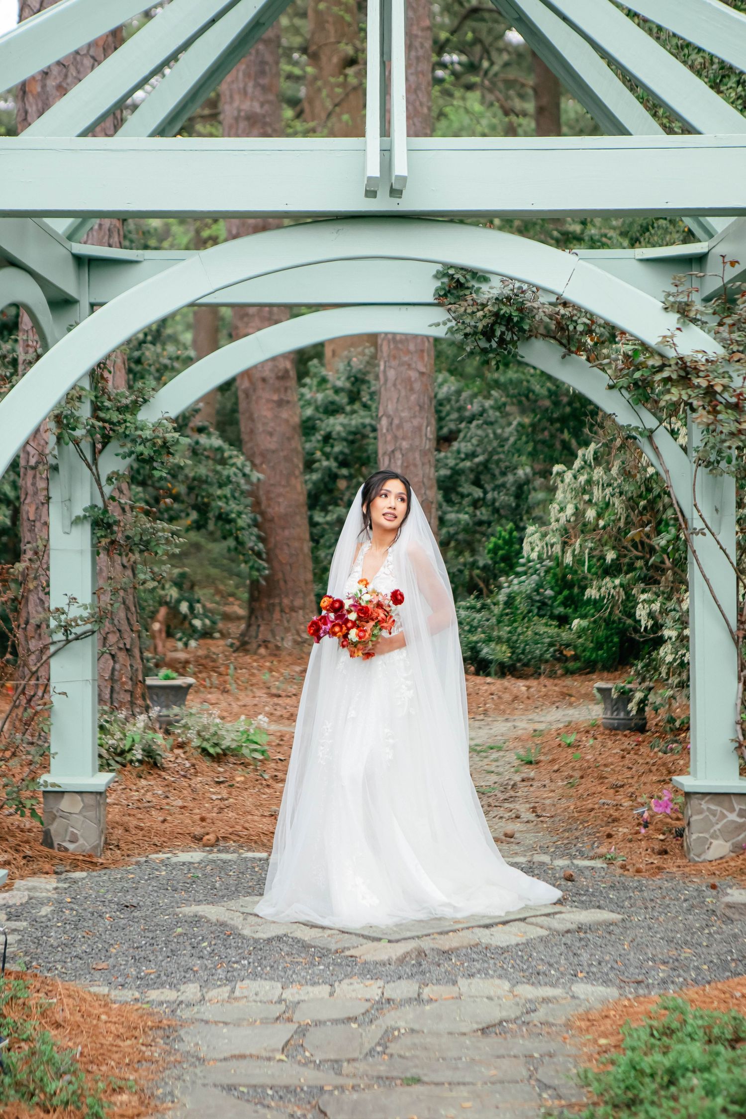 Bride in white gown holds red bouquet under mint green garden arch.