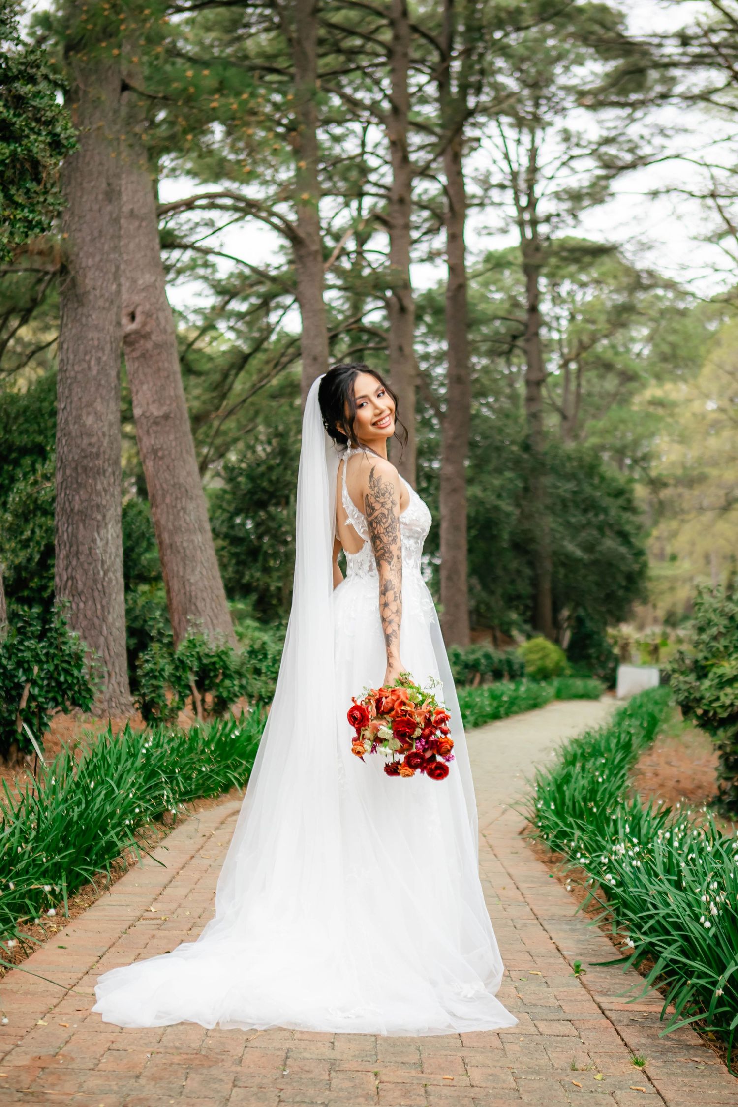 Bride in lace gown holds red bouquet on garden path lined with tall pines.