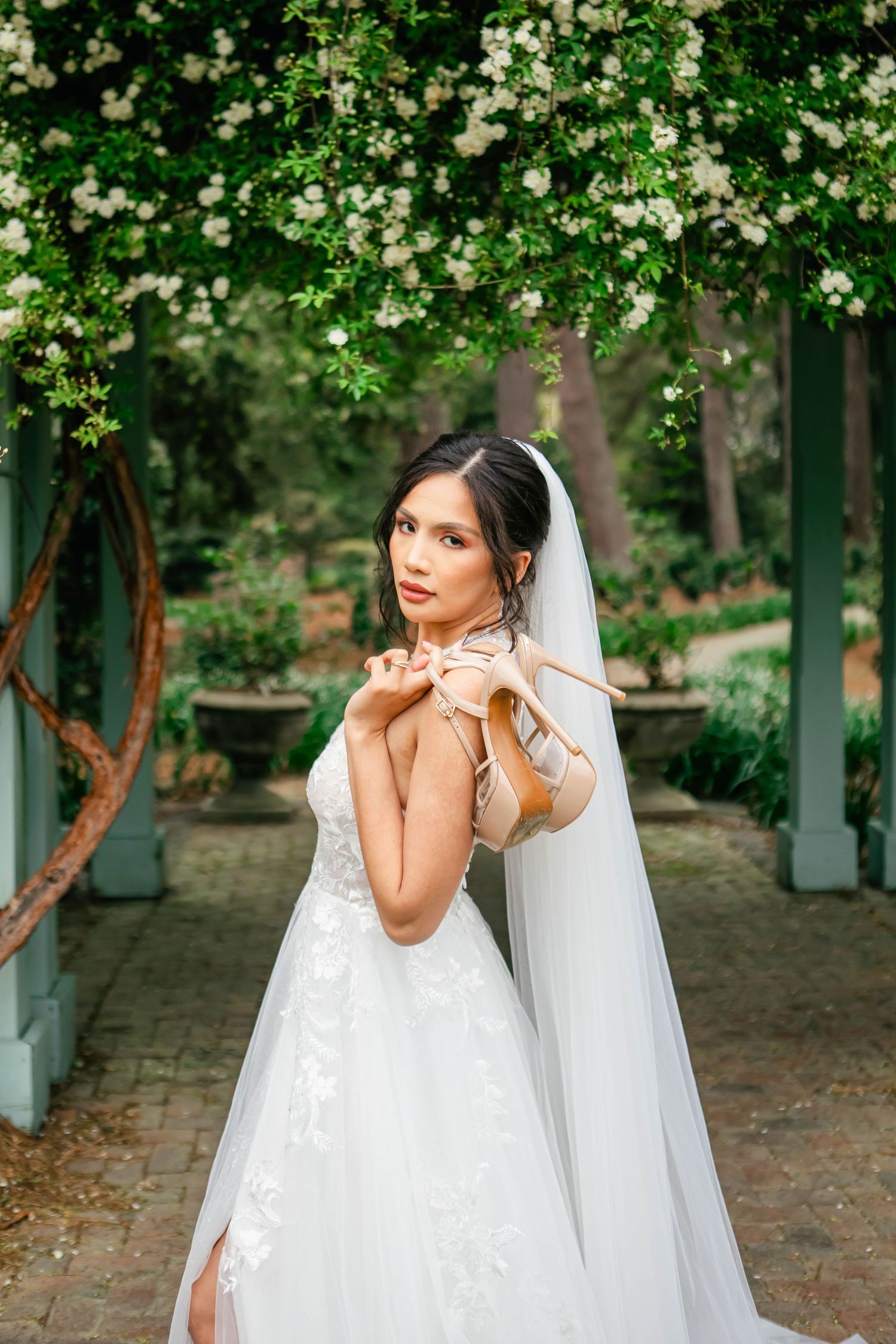 A bride in a white lace wedding dress poses elegantly among green trees and foliage.