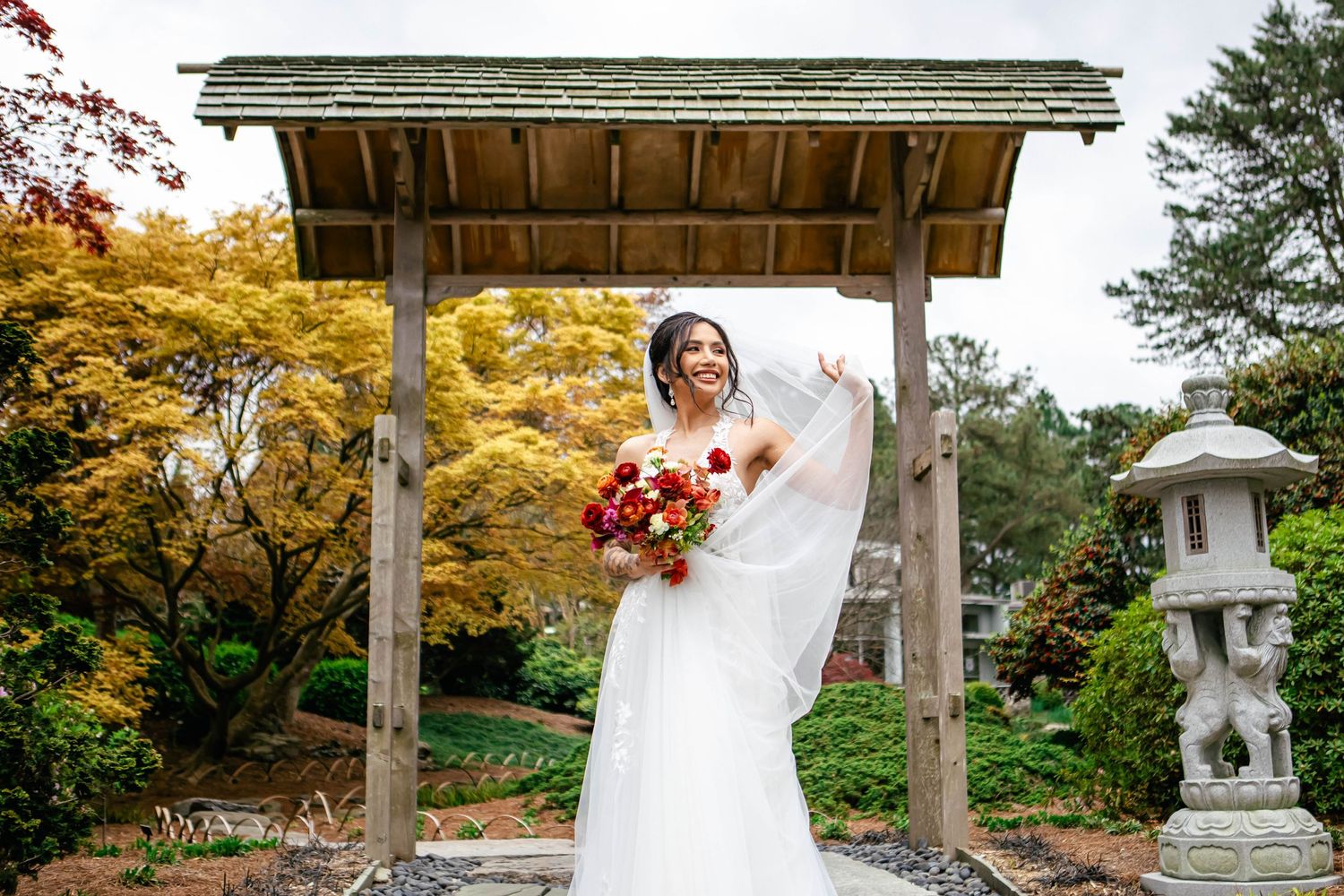 Bride with red bouquet stands under wooden pavilion with autumn foliage.