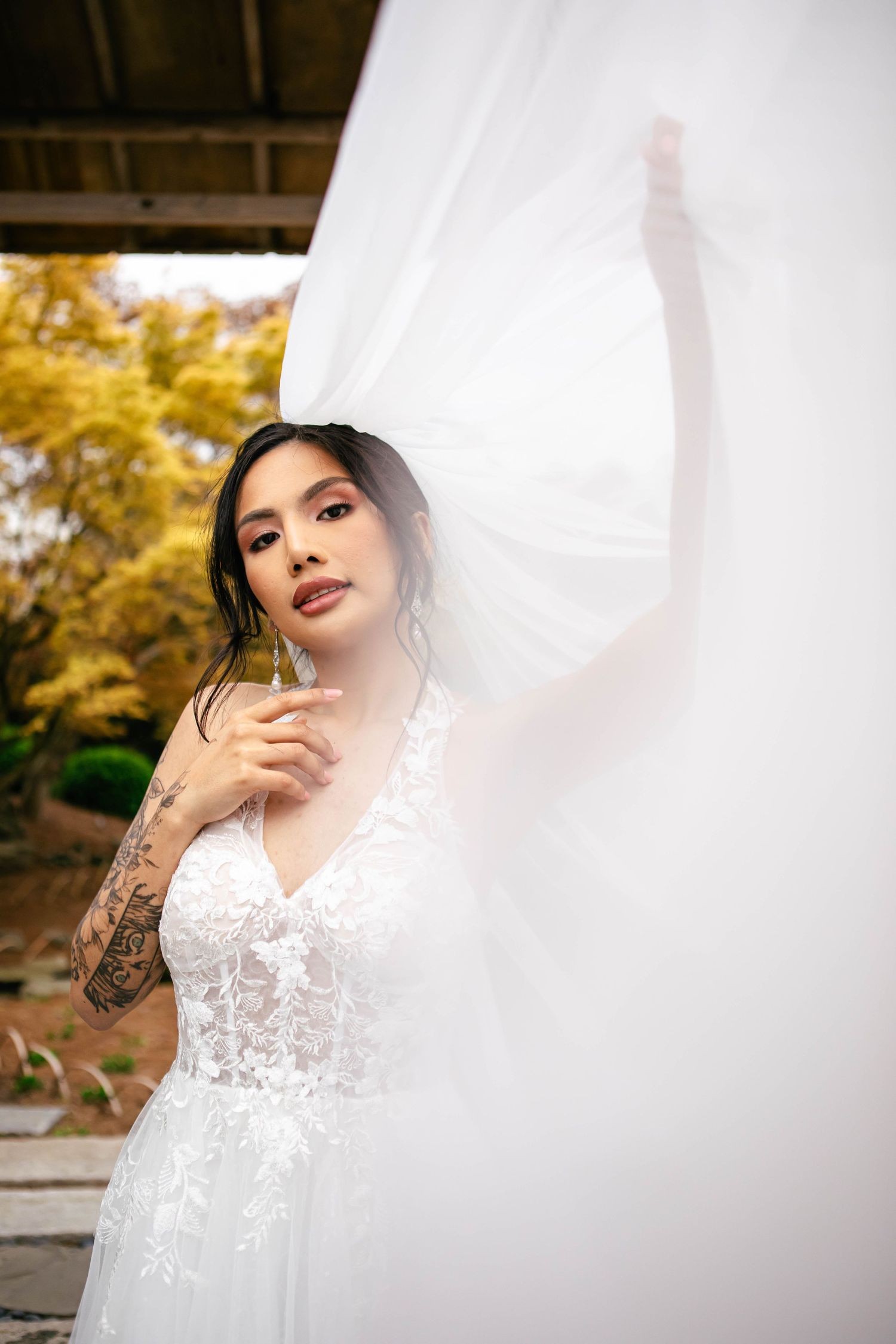 A bride in a white lace dress poses against a sheer veil with autumn foliage in the background.