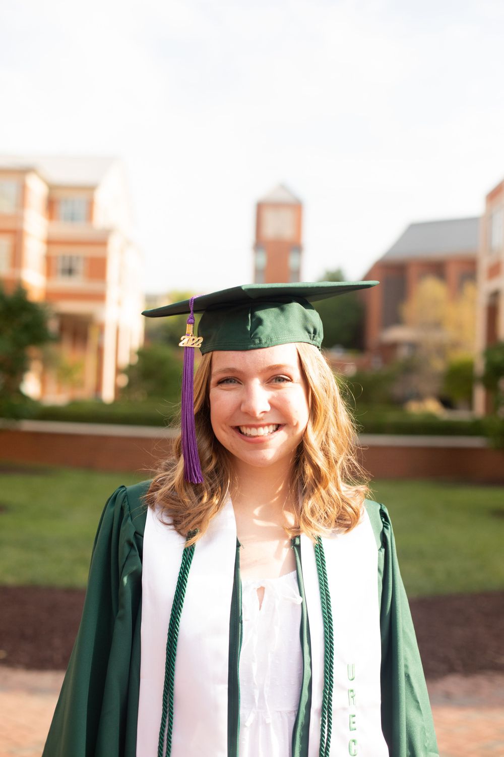 Graduate in green regalia and cap smiles brightly on campus grounds.