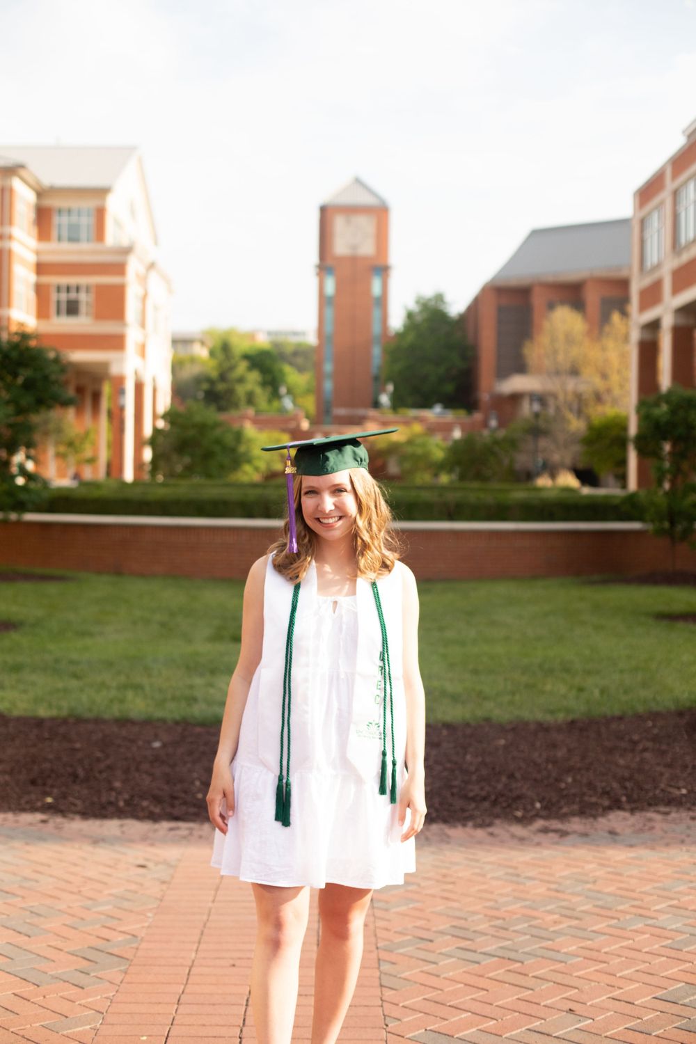 Graduate in white dress and green stole stands on brick path with campus clock tower behind.
