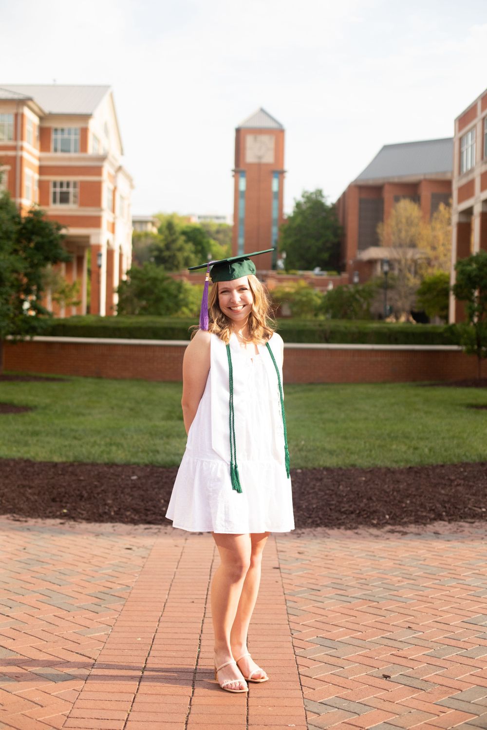 Graduate poses in white dress and green regalia on brick courtyard with clock tower visible.