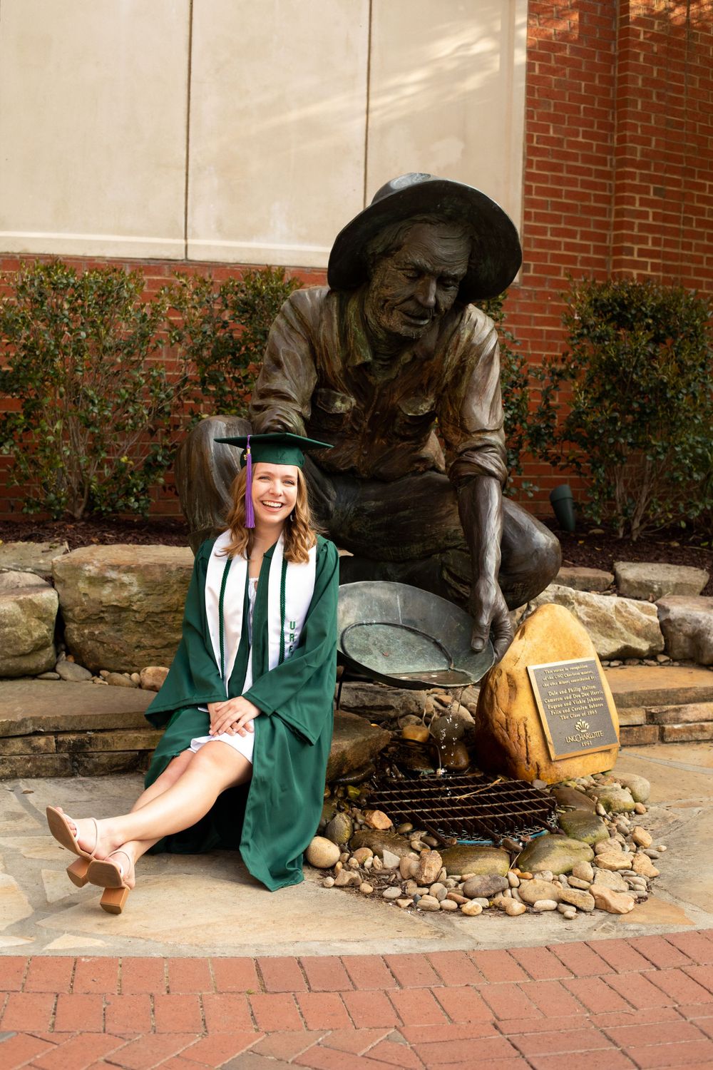 Graduate in green regalia poses with bronze statue outside campus building.