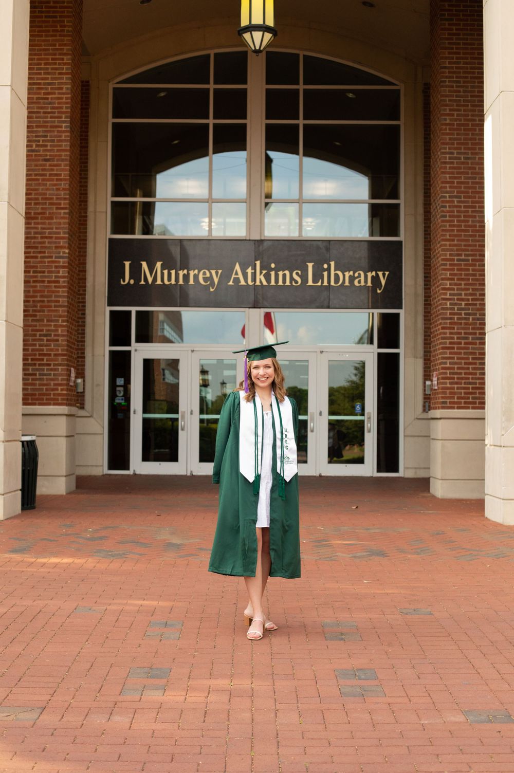 Graduate stands in green regalia outside J. Murray Atkins Library entrance.