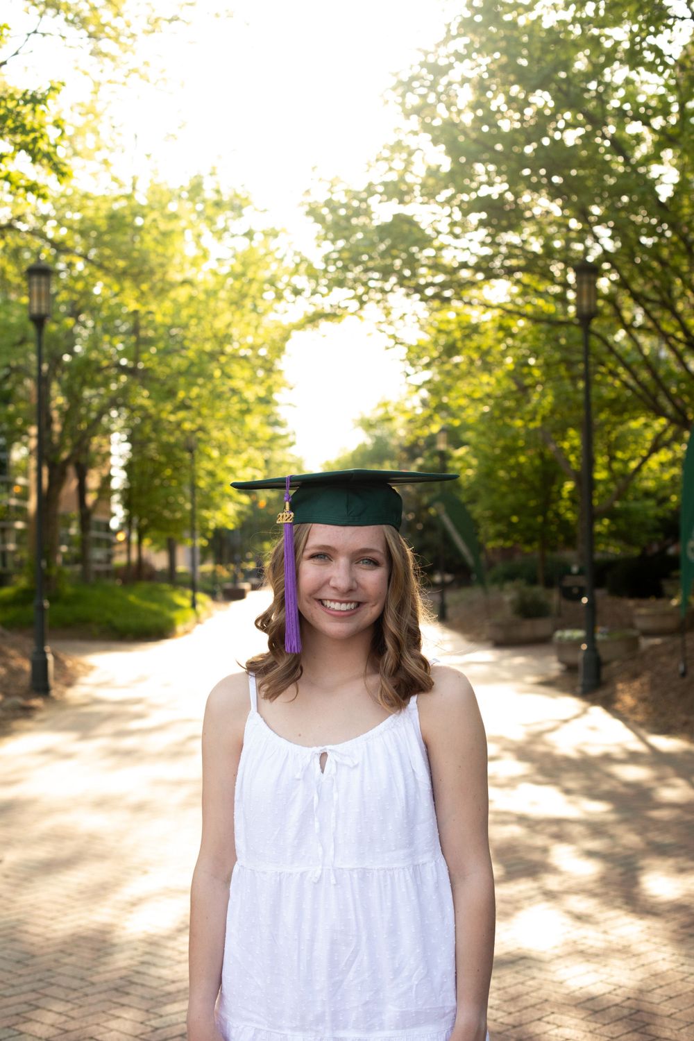 Graduate in white dress and cap poses on a tree-lined brick pathway during golden hour.