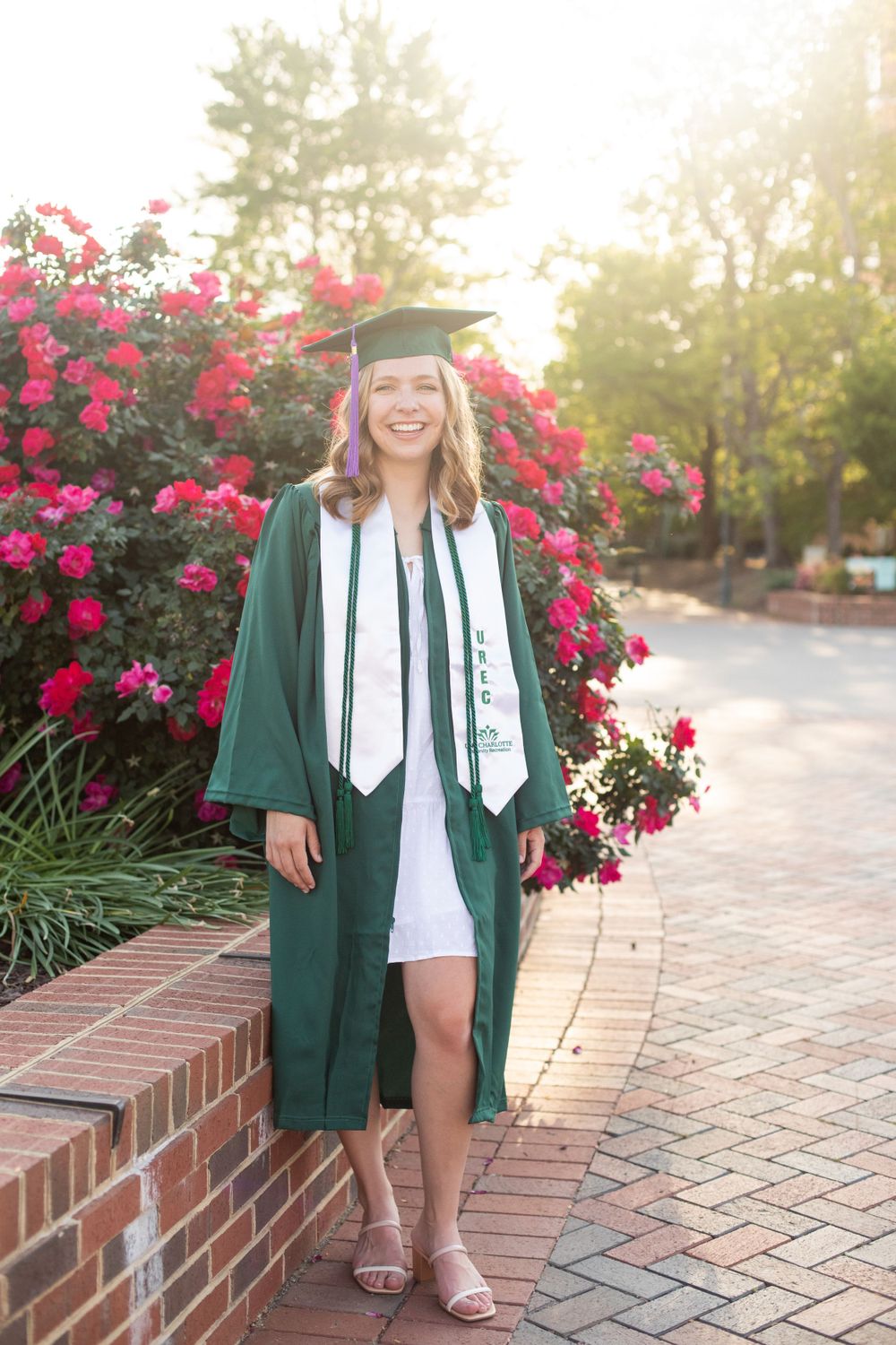 Graduate wearing green gown and white stole poses near blooming red flowers and brick wall.
