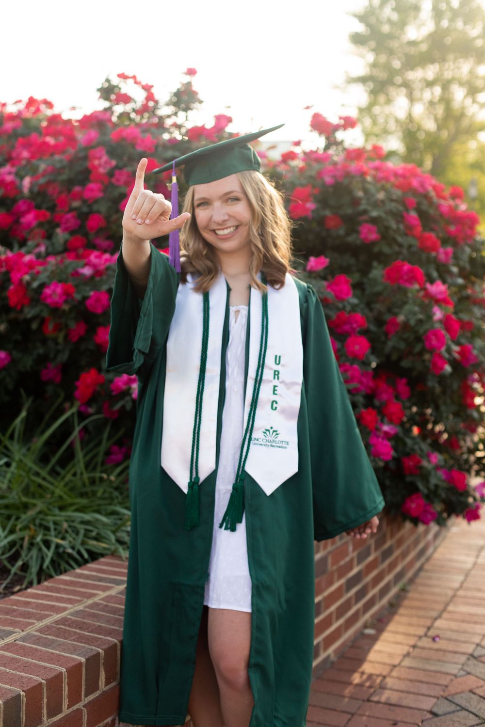 Graduate in green cap and gown gives thumbs up near red flowering bushes.