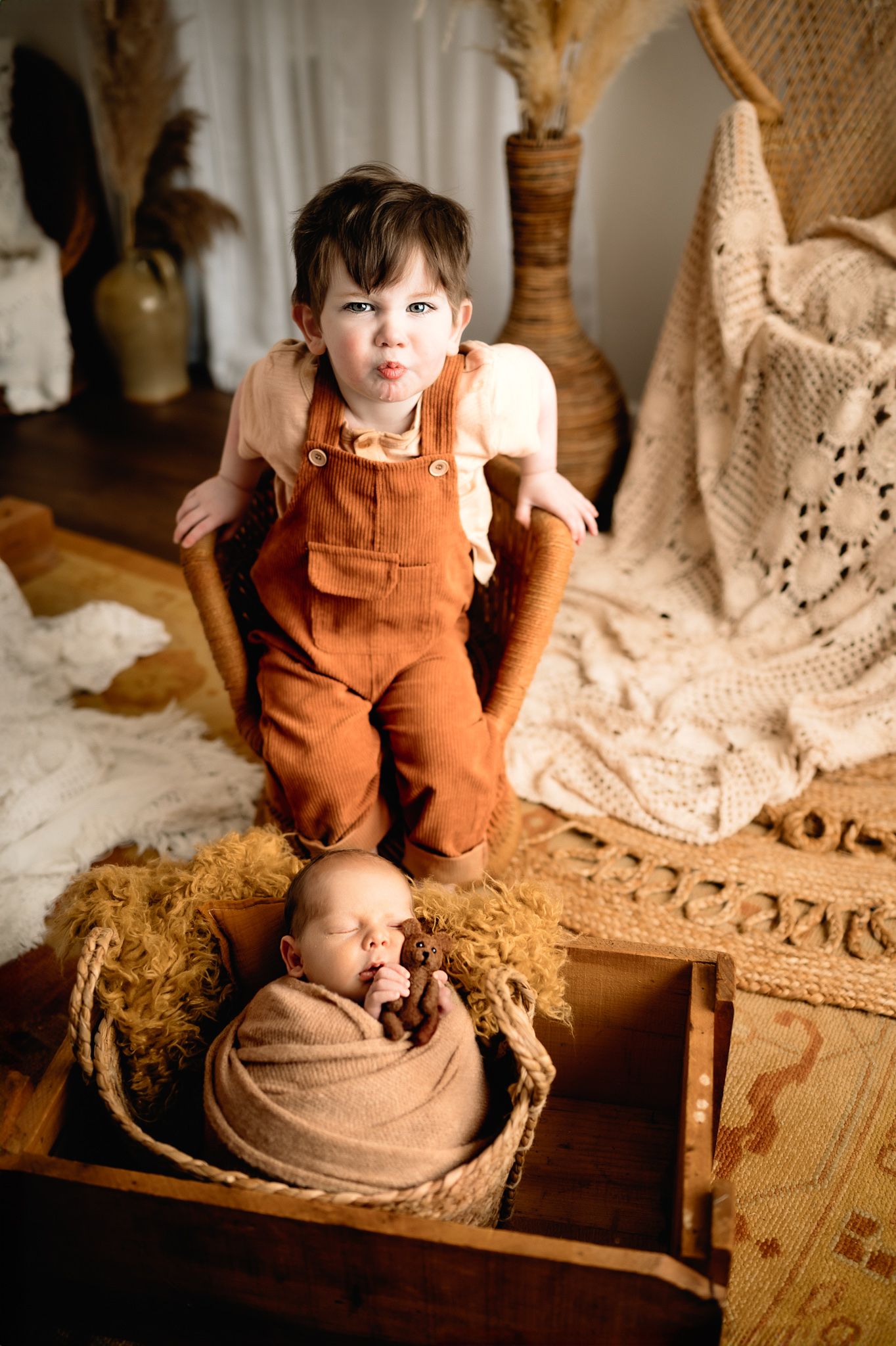 Child in orange overalls standing near a newborn baby in vintage-styled setting.