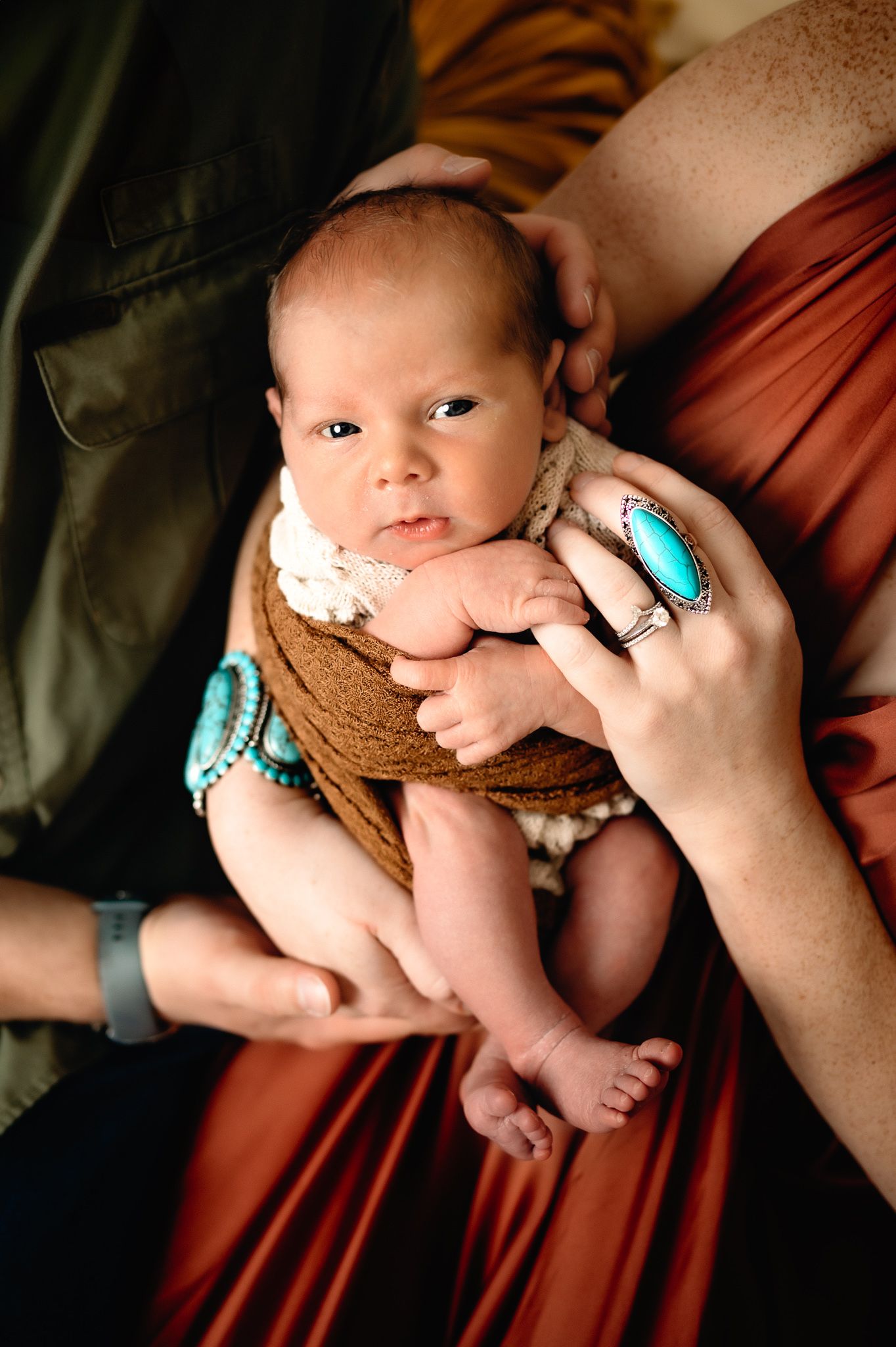 Newborn baby wearing brown wrap with turquoise jewelry details in indoor setting.