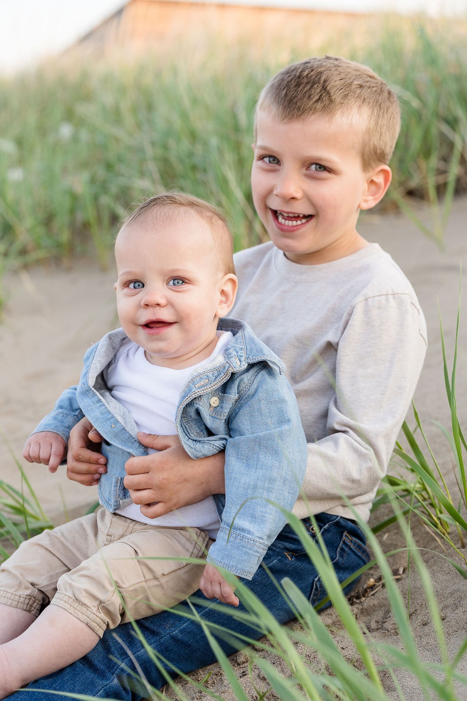 Two young brothers sitting on sandy beach dunes, older boy smiling and hugging baby sibling dressed in denim jacket.