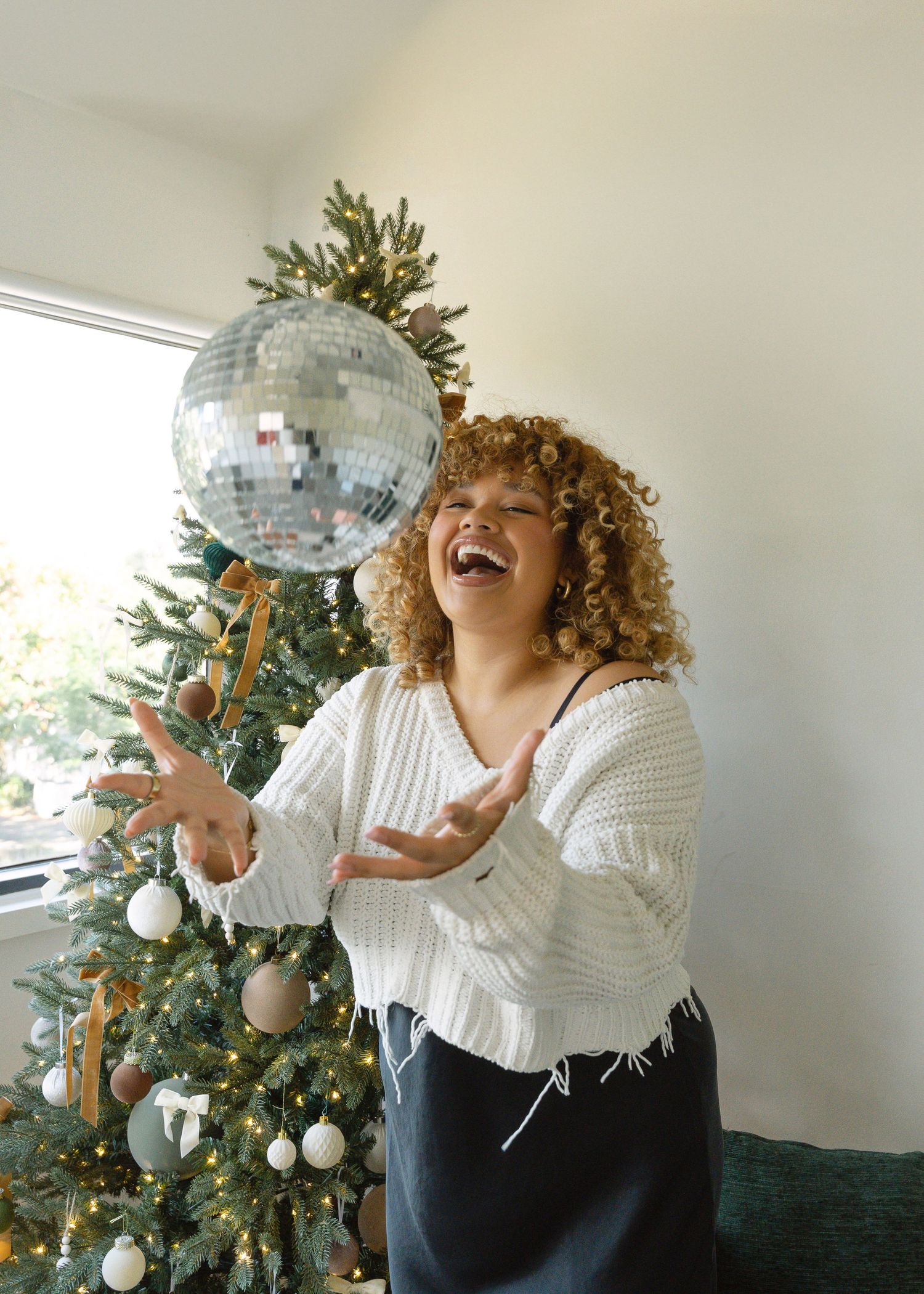 Mother in white outfit embraces young child beside decorated Christmas tree with wrapped gifts.