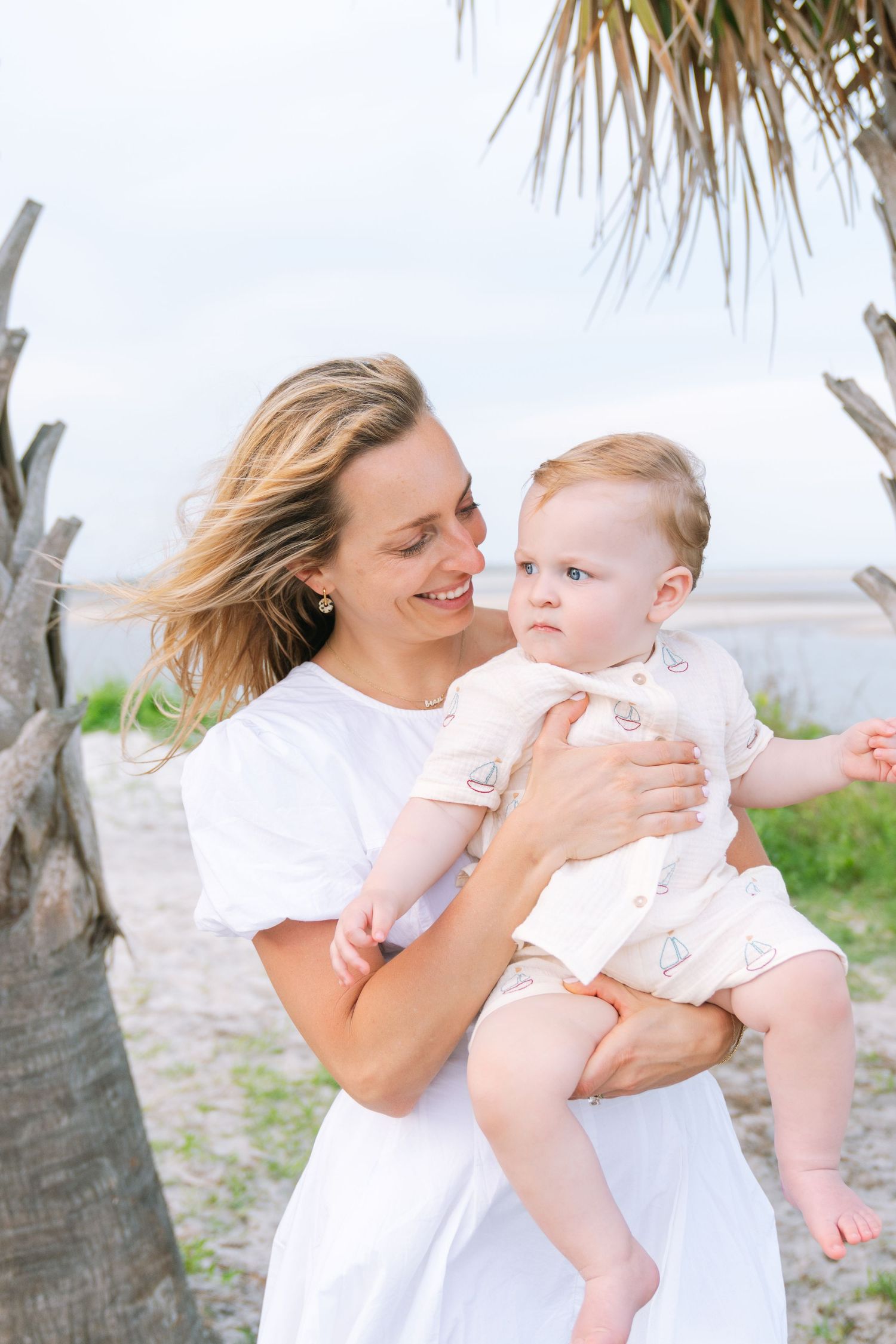 Mother in a white dress holding her baby beside a palm tree during an outdoor family photography session in the fall.