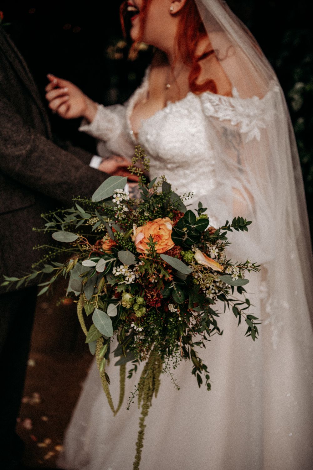A wedding bouquet featuring orange roses, eucalyptus, and trailing greenery captured in a moody, dark setting.