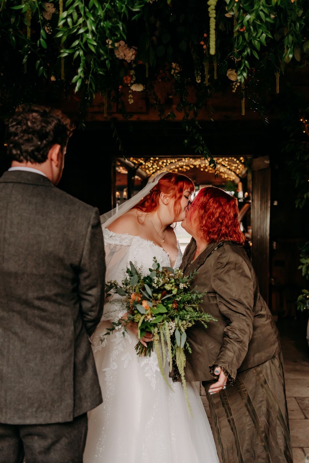 Two people in wedding attire share an intimate moment under twinkling lights with a floral bouquet and greenery.