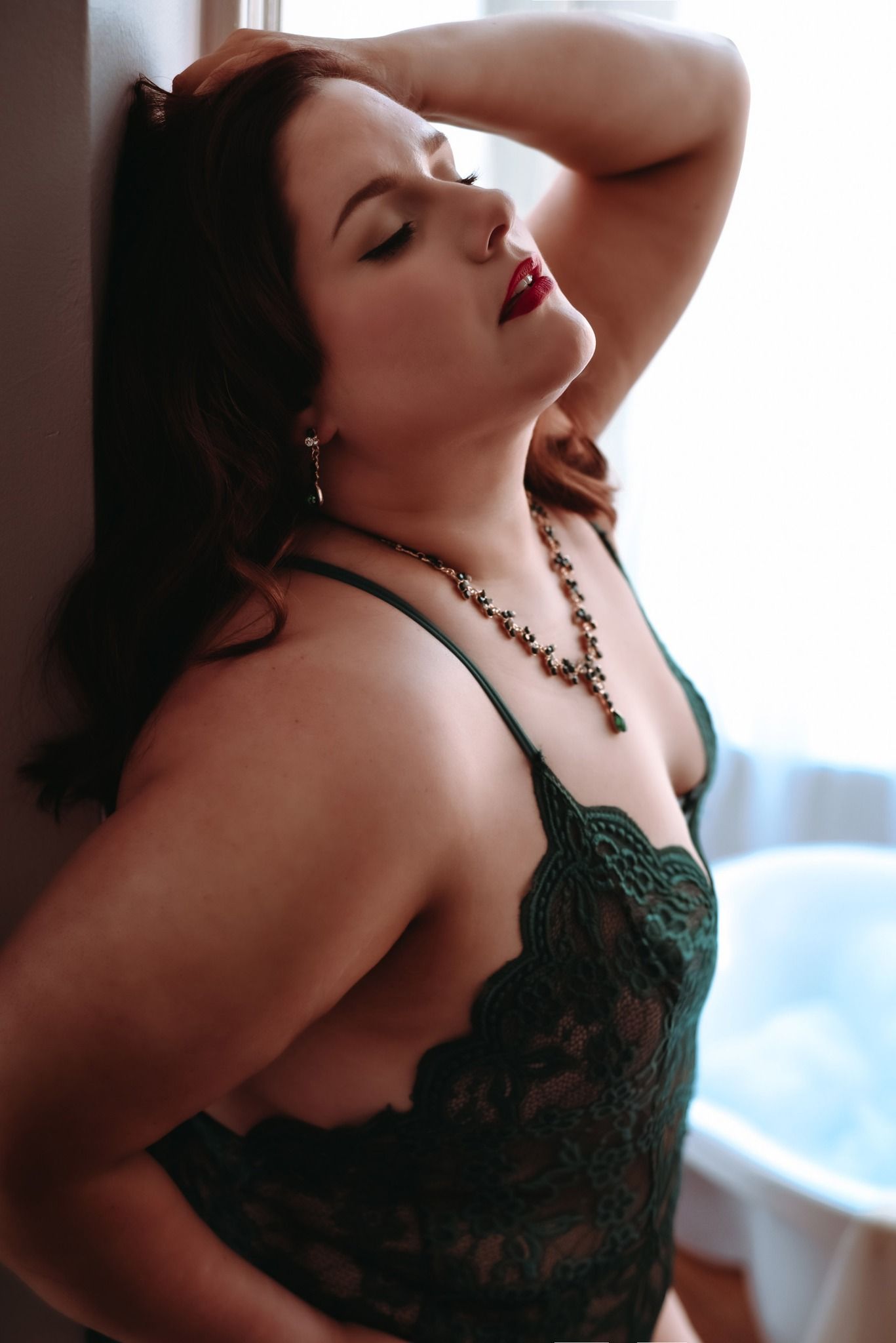 Profile portrait showing dark lace lingerie with necklace in soft moody lighting against light background.