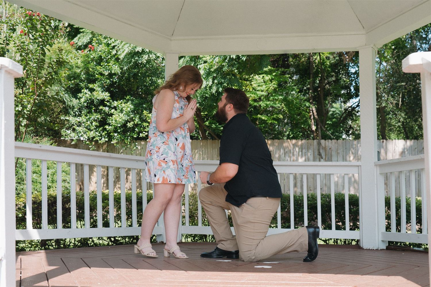 A romantic proposal takes place in a white gazebo surrounded by lush greenery on a sunny day.
