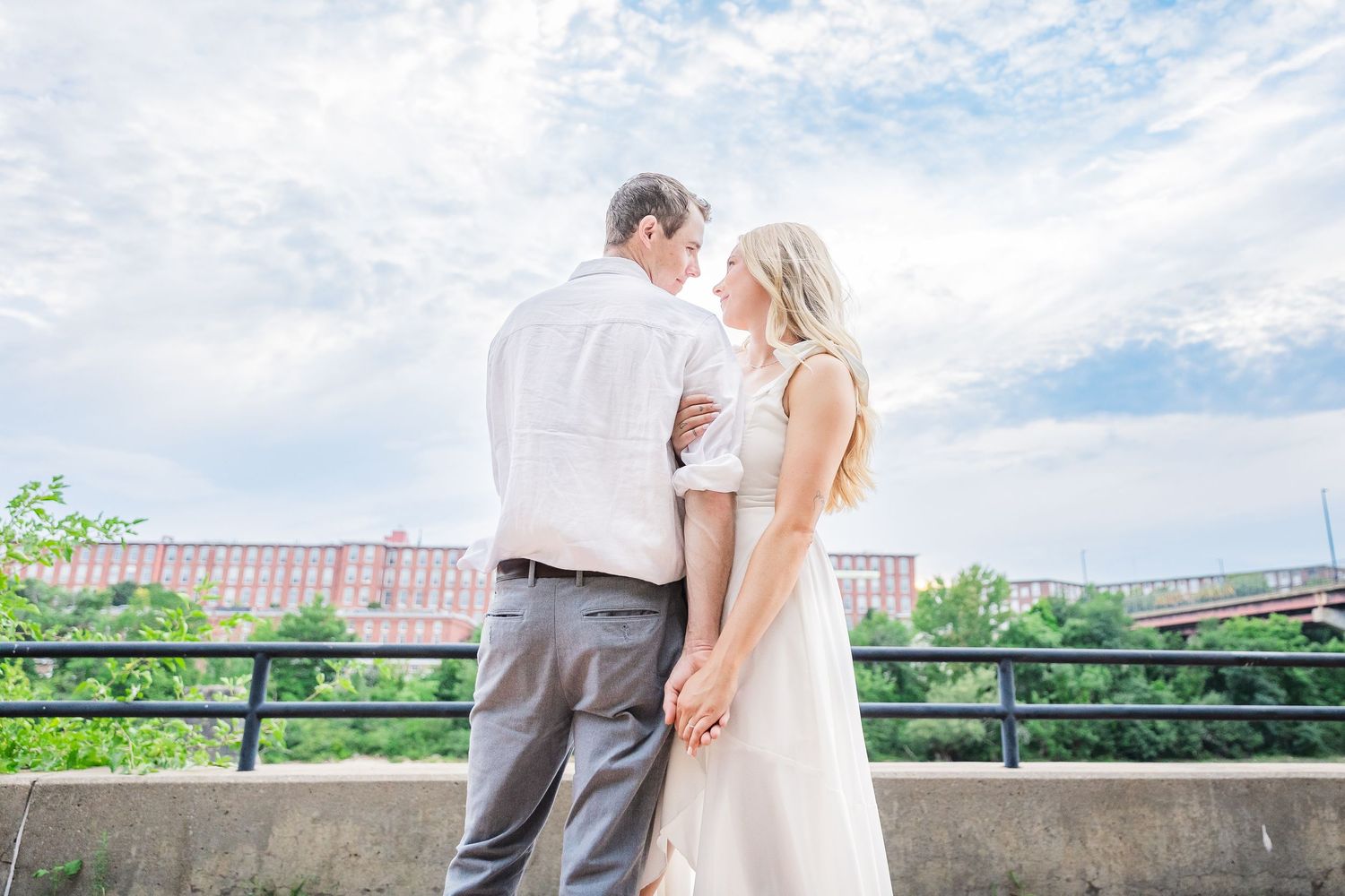 Couple embraces in front of industrial brick building with blue sky backdrop.
