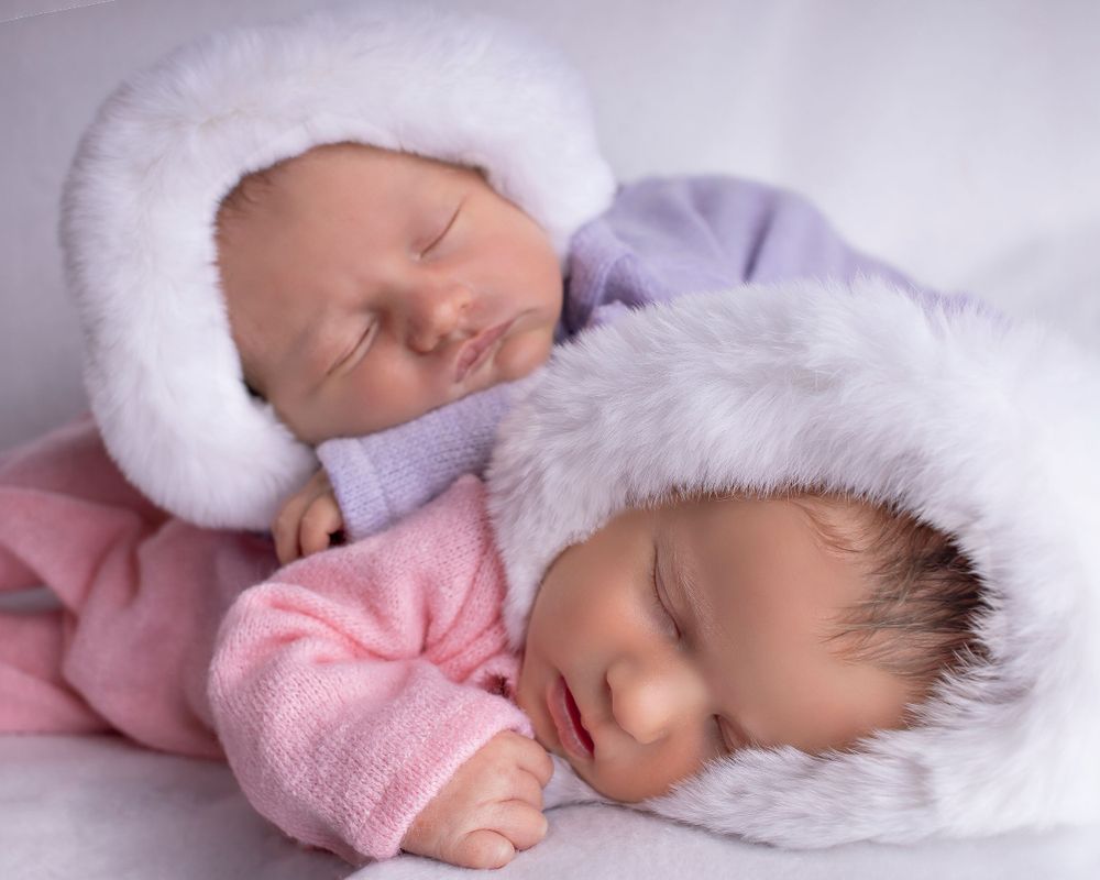 Twin newborn girls sleep soundly while cuddling in furry bonnets