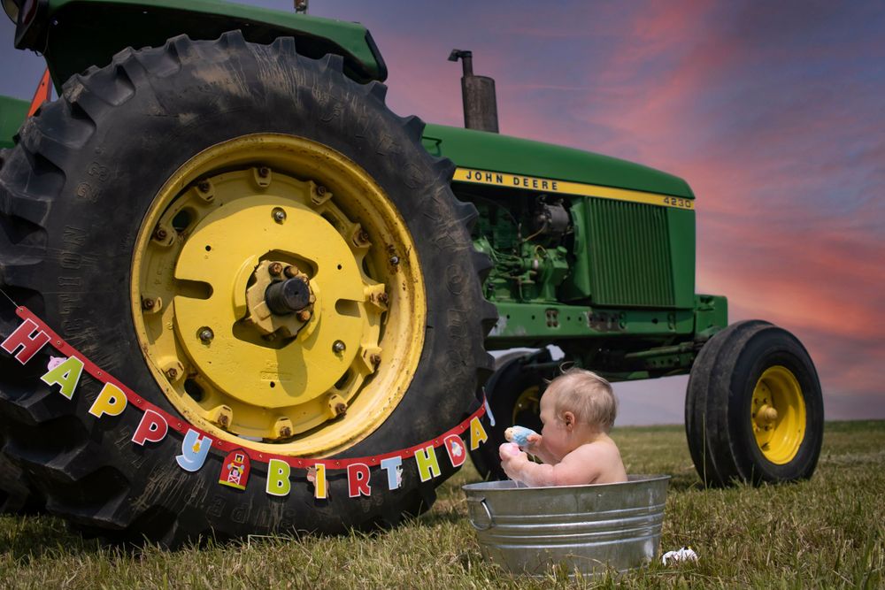A baby boy has his back to the camera while taking a bubble bath in a bucket. A tractor and dramatic sky are in the background
