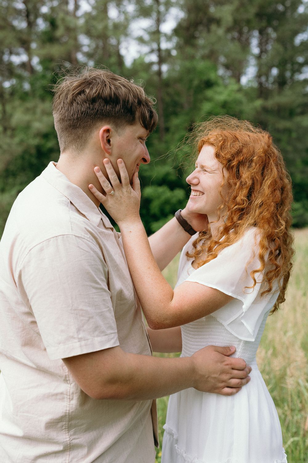 A romantic couple in white outfits shares tender moments in a sunny meadow with pine trees in the background. wedding photographer