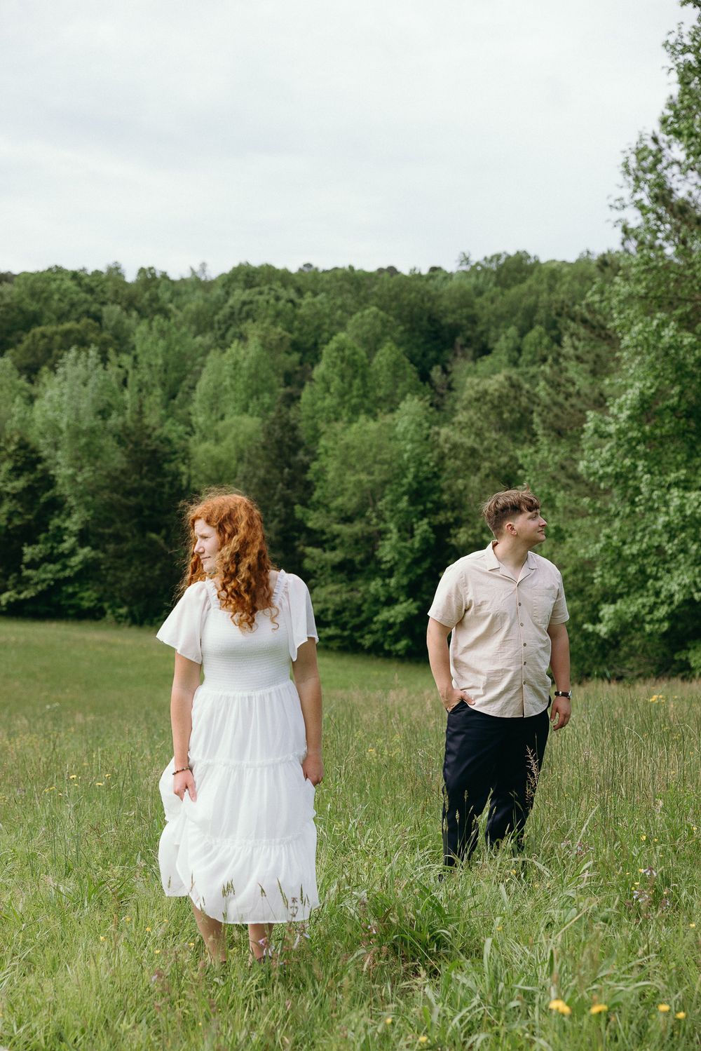 A couple in white clothing walks through a lush summer meadow surrounded by trees. Le Château du Cœur Wedding Photographer