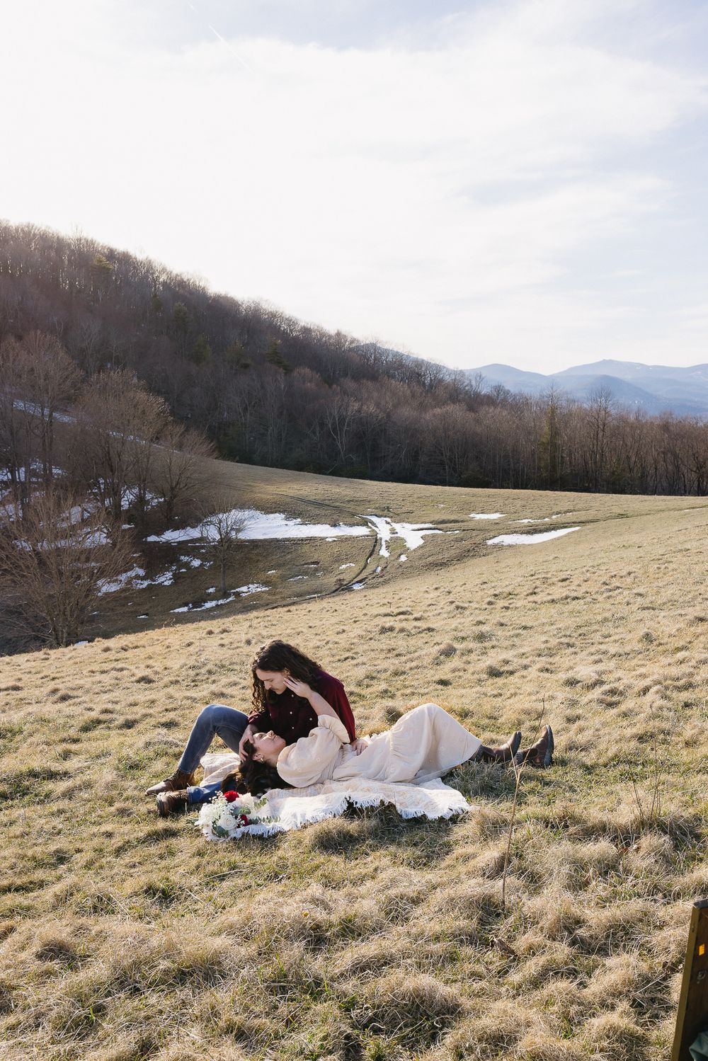 A couple lies together on a grassy hillside with mountains and patches of snow visible in the distance. Castle  Ladyhawke Wedding Photograph
