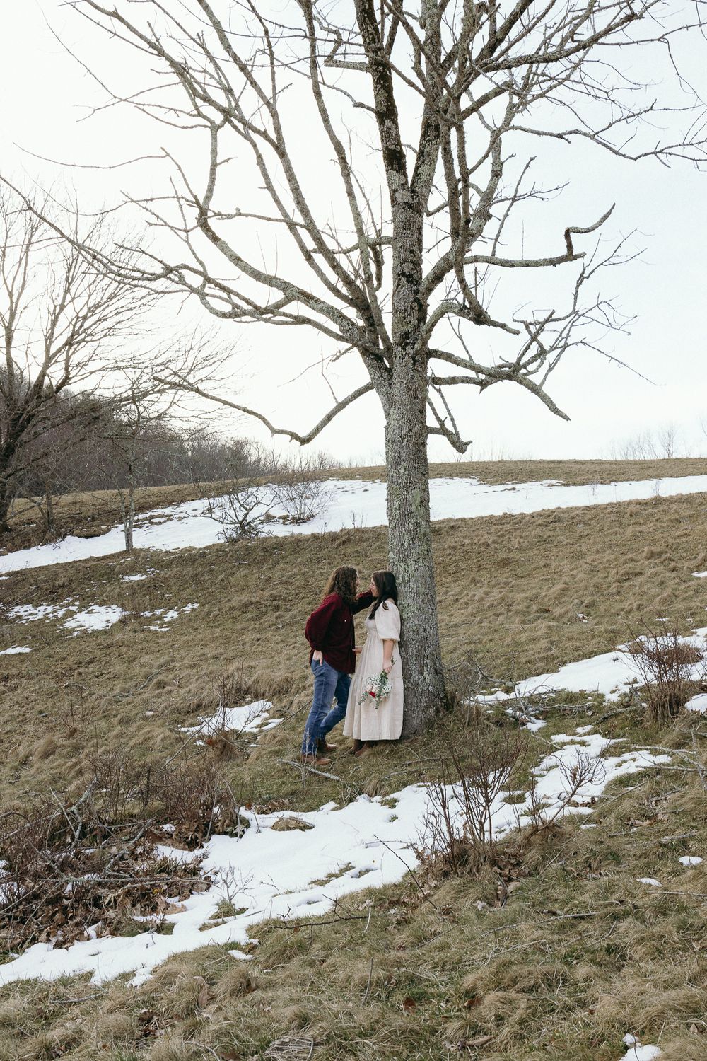 A couple walks along a snowy hillside with bare winter trees and mountains in the distance. Twickenham House Wedding Photographer