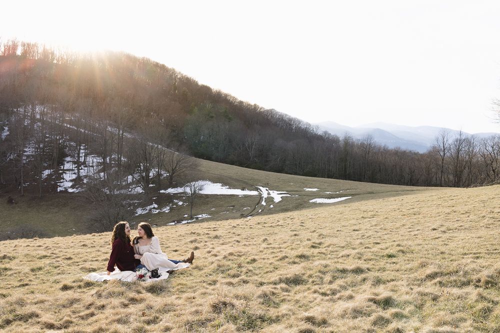A couple sits on a grassy hillside overlooking a scenic mountain landscape at sunset. Twickenham House Wedding Photographer
