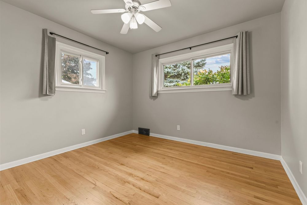Sunlit bedroom featuring hardwood floors, ceiling fan, and two windows with gray curtains.